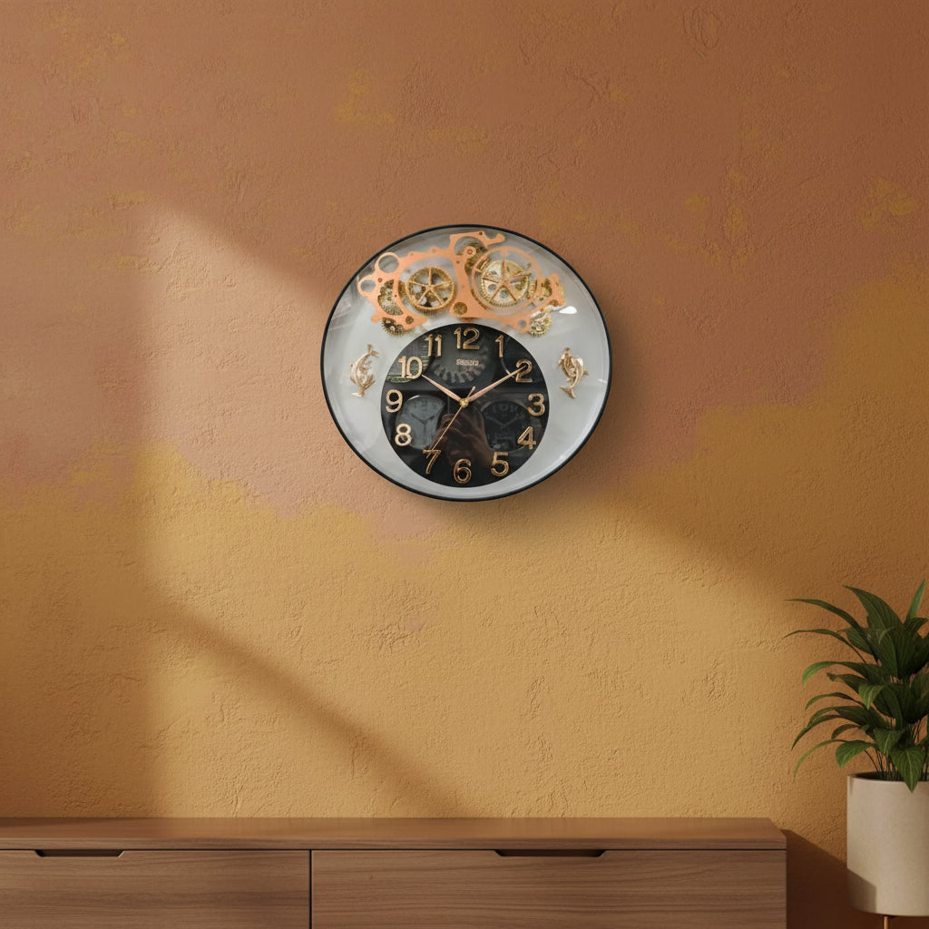 A round wall clock with a black central face, gold Arabic numerals, and a decorative upper section showing gold-toned gear and wheel motifs against a clear outer ring.