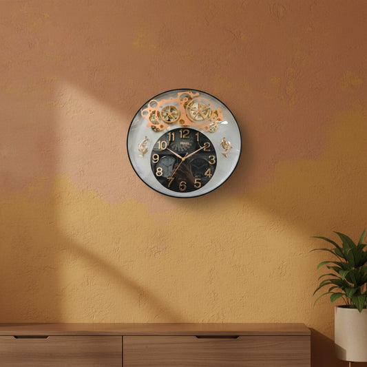 A round wall clock with a black central face, gold Arabic numerals, and a decorative upper section showing gold-toned gear and wheel motifs against a clear outer ring.